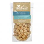 A glossy pouch of Joe & Seph’s Vegan Salted Caramel Popcorn sits against a pale backdrop. Warm amber caramel coats irregularly popped kernels, showing glistening crystals of salt. The brand logo appears center on the bag, with vegan labeling and golden accents. Soft matte backdrop and soft lighting.