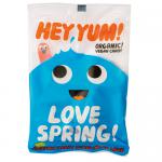 A bright photo shows several HEY YUM! Love Spring organic sweets packets arranged on white. Pastel floral graphics and playful typography decorate glossy wrappers. Small clear windows reveal tiny round candies in soft pink, mint and lemon hues. A green organic label sits near the bold logo. Upfront.