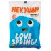 A bright photo shows several HEY YUM! Love Spring organic sweets packets arranged on white. Pastel floral graphics and playful typography decorate glossy wrappers. Small clear windows reveal tiny round candies in soft pink, mint and lemon hues. A green organic label sits near the bold logo. Upfront.