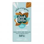 Photograph shows a dark chocolate bar with glossy squares, threaded ribbons of golden caramel and scattered coarse sea salt crystals. Mixed seeds — pumpkin, sunflower, sesame, flax — dot the surface. Wrapping with rustic, kraft-style label and natural tones frames the bar. Soft light reveals texture