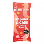 Bright red-orange bag of Brave Roasted Peas Paprika & Chilli sits on a wooden surface. Crisp roasted green peas tumble from a torn corner beside scattered paprika powder and red chili slices. Bold typography and spice illustrations decorate the front against a warm gradient backdrop. Soft studio-lit