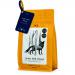 A matte black bag sits against a textured wooden backdrop, its label depicting moonlit trees. Rich, glossy coffee beans spill forward in a small pile, catching soft light that highlights their oil sheen. Muted tones and cool shadows evoke a quiet, shadowed forest. A faint steam curls above the beans