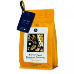 A matte black coffee pouch stands upright on a wooden surface, its kraft paper label reading Dark Woods Common Grounds Barrel Aged. A small barrel illustration accents the label. Loose coffee beans spill at the base. Warm, directional light and subtle shadows create a rustic, artisanal mood. Nearby.
