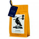 A matte black coffee bag stands upright on a wooden surface. A stark silhouette of a crow perched on a gnarled tree dominates the cream label, with bold serif Dark Woods text beneath. Amber-brown coffee beans scatter at the base; 250g appears in small type. A subtle matte sheen and creased top seal.