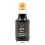 Photo shows six slender glass bottles of smoked soy sauce arranged in two neat rows on a neutral background. Each 150ml bottle holds dark amber liquid, displays a monochrome label bearing The Wasabi Company logo, and is topped with a silver screw cap. Soft studio lighting creates gentle reflections.