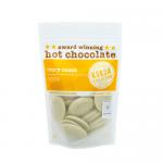 A glossy tin of Kokoa Collection White Hot Chocolate rests on a warm wooden table. The ivory label shows delicate cocoa pod illustrations, brown serif lettering and a small Ivory Coast emblem. A porcelain cup with rising steam sits beside it in soft, natural light. Light highlights cream tones, warm.