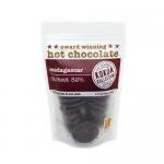 A matte-black tin labeled Kokoa Collection Organic Darkest Hot Chocolate Madagascar 82% sits on a rustic wooden table. Gold trim and minimalist white type contrast rich cocoa illustrations. Scattered cocoa nibs and a dusting of deep brown powder frame the container. Soft light accents matte details.
