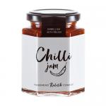 Six glass jars line a rustic wooden board, filled with glossy red chilli jam and varied brown chutneys. Each jar wears a cream label with Hawkshead Relish text and a black lid. Soft natural light highlights jar textures and casts subtle reflections and shadows on the surface. A small brass spoon lies