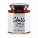 Six glass jars line a rustic wooden board, filled with glossy red chilli jam and varied brown chutneys. Each jar wears a cream label with Hawkshead Relish text and a black lid. Soft natural light highlights jar textures and casts subtle reflections and shadows on the surface. A small brass spoon lies