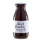 Six glass jars of Hawkshead Relish Black Garlic Ketchup sit in a tidy row on a wooden board, their dark, glossy contents visible through clear glass. Each jar bears a simple cream label with black typography and garlic illustration; scattered chutney jars and spoons frame the scene in soft daylight.