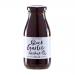 Six glass jars of Hawkshead Relish Black Garlic Ketchup sit in a tidy row on a wooden board, their dark, glossy contents visible through clear glass. Each jar bears a simple cream label with black typography and garlic illustration; scattered chutney jars and spoons frame the scene in soft daylight.