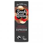 A slender dark chocolate mini bar rests on a pale surface. Matte wrapper in deep brown and cream shows subtle espresso imagery. One corner is broken to reveal a glossy, dense interior with fine texture and a few coffee specks, clean edges and a compact bite-sized shape. Soft warm light adds depths.