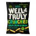 A glossy green-and-white pouch of Well&Truly Crunchy Sour Cream & Onion stands upright. It displays a bold logo, the flavor name, and an image of ridged crisps topped with chives. Crinkles along the edges reflect studio light; icons and a nutrition panel sit on the back. A small tag appears atop.