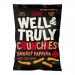 A glossy red-orange bag displays the Well&Truly logo across the top. Smoky paprika graphics and scattered crunchy pieces decorate the front. A clear window reveals textured crisps. Nutritional information and a resealable zip appear along the top edge. Bright yellow accents and a smaller flame icon.