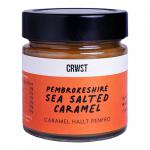 Six glass jars of CRWST Pembrokeshire Sea Salted Caramel are arranged in two rows on a neutral surface. Rich amber sauce shows through the jars. Cream labels with black lettering and coastal motifs and metal lids reflect soft studio light.