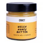 A row of six glass jars shows CRWST Welsh Honey Butter and CRWST Dessert Sauces. Each jar features a cream-colored label with black and gold lettering, honeycomb motifs and a screw-top lid. Thick, amber-brown contents glisten through clear glass under soft studio lighting. Small bee icon shows up.