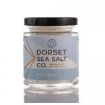 Six clear 100g glass jars with silver lids sit in a neat grid. Each jar shows coarse white sea salt visible through the glass and features a simple cream label reading Dorset Sea Salt in understated type. Soft natural light highlights the crystalline texture. Lit shadows cross a pale linen backdrop.