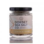 A cluster of six small glass jars with metal screw lids sits on a pale surface. Each jar displays a bright lemon and thyme label and shows coarse off-white salt flecked with tiny lemon zest and dried green thyme fragments. Soft natural light highlights the texture. A soft shadow falls nearby. Seaside