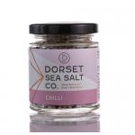 A group of six clear 100g glass jars of Dorset chilli sea salt sits on a neutral surface. Each jar displays a white label with bold black text and a small red accent. Coarse pale salt shows scattered red chilli flakes. Soft studio lighting creates gentle reflections on the glass. tiny coastal icons.