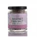 A group of six clear 100g glass jars of Dorset chilli sea salt sits on a neutral surface. Each jar displays a white label with bold black text and a small red accent. Coarse pale salt shows scattered red chilli flakes. Soft studio lighting creates gentle reflections on the glass. tiny coastal icons.