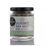 A clear 100g glass jar with a silver screw lid contains white, flaky sea salt speckled with green celery pieces. The label displays Dorset Sea Salt branding with green accents and a small celery illustration; the jar sits on a neutral surface.