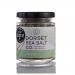 A clear 100g glass jar with a silver screw lid contains white, flaky sea salt speckled with green celery pieces. The label displays Dorset Sea Salt branding with green accents and a small celery illustration; the jar sits on a neutral surface.