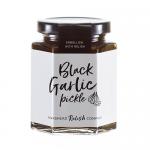 Six glass jars sit in a neat row, each bearing a Hawkshead Relish label. One jar reveals glossy, deep-brown black garlic pickle; others show textured chutneys and condiments in amber, ruby and green hues. Labels, metal lids and a simple wooden backdrop complete the scene. Warm light catches jar rims
