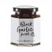 Six glass jars sit in a neat row, each bearing a Hawkshead Relish label. One jar reveals glossy, deep-brown black garlic pickle; others show textured chutneys and condiments in amber, ruby and green hues. Labels, metal lids and a simple wooden backdrop complete the scene. Warm light catches jar rims