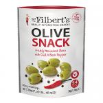 Glossy green olives fill a small clear jar, their skins speckled with red chilli flakes and cracked black pepper. A simple label reads Mr Filberts. The jar sits on a wooden board beside scattered olives and peppercorns, with warm natural light highlighting oil sheen. Tiny oil drops catch light, now.
