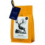 A matte, dark-brown bag branded Dark Woods displays a stylized deer silhouette before a moonlit pine forest. Copper lettering names Deer Hill Ground Coffee 250 g. The pouch sits on weathered wood alongside scattered roasted beans under warm, moody studio lighting. A faint steam rises. Soft shadowing