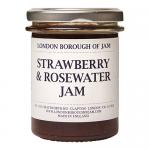 Six glass jars of strawberry and rosewater jam sit arranged in a tidy group, showing a soft pink-red preserve. Cream labels feature elegant script and rose illustrations; gold lids catch light. The jam’s glossy texture reveals tiny fruit pieces.