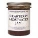 Six glass jars of strawberry and rosewater jam sit arranged in a tidy group, showing a soft pink-red preserve. Cream labels feature elegant script and rose illustrations; gold lids catch light. The jam’s glossy texture reveals tiny fruit pieces.
