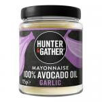 A glossy jar of Hunter & Gather garlic avocado oil mayonnaise sits center, pale creamy spread visible through clear glass, green-and-white label with brand logo. Surrounding it are six small condiment tubs with colorful lids and matching labels, arranged on a white surface.