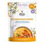 A bright orange sea buckthorn powder sits in a clear glass jar beside a labeled pouch reading Arctic Power Berries. Fresh orange berries and green leaves scatter around a wooden scoop on a clean white background in a minimalist lifestyle arrangement.