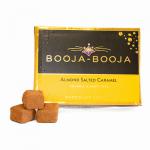 Close-up of a small rectangular box of Booja-Booja Almond Salted Caramel truffles. Matte brown and cream packaging shows elegant typography and an almond-splash graphic. Eight glossy, round truffles sit beside the box, their caramel centers hinting through slight cracks in the chocolate shells.