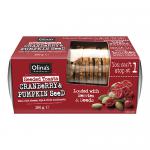 A clear plastic pack displays golden-brown rectangular seeded toasts studded with glossy dried cranberries and green pumpkin seeds. Each slice shows a rustic, grain-speckled texture. A simple bakery sleeve with green accents labels the stacked loaves.