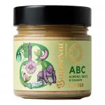 A glass jar with a cream-colored lid reveals a smooth, pale beige nut butter. The label shows illustrations of almonds, Brazil nuts and cashews with elegant typography and muted tones. A spoon beside the jar lifts a glossy, velvety swirl of the spread. Whole nut halves scatter on a warm wooden slab.