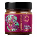 A glass jar of creamy cacao and hazelnut butter stands center on a neutral surface. The warm brown spread shows a glossy, smooth surface; the label displays brand text and cocoa imagery. A wooden spoon rests beside it with a small swirl of spread. Soft natural light brings out glossy texture on oak.