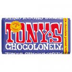 A rectangular Tony's Chocolonely bar in branded wrapper sits broken into chunky, uneven squares. Dark-milk chocolate shows a glossy surface and dense texture, with visible salted pretzel fragments and golden toffee shards embedded across the pieces, scattered crumbs on the surface. Packaging nearby.