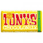 Bright, unevenly segmented Tony's Chocolonely wrapper dominates the frame, showing bold white logo across warm brown and orange background. Partly unwrapped milk chocolate bar reveals almond pieces and nougat layers; crumbs and a honey sheen accent textures on a simple surface. Warm glow emphasizes.