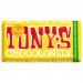 Bright, unevenly segmented Tony's Chocolonely wrapper dominates the frame, showing bold white logo across warm brown and orange background. Partly unwrapped milk chocolate bar reveals almond pieces and nougat layers; crumbs and a honey sheen accent textures on a simple surface. Warm glow emphasizes.