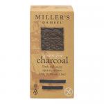 Image shows a bakery scene: six charcoal-gray round biscuits nest on crumpled kraft paper beside a simple cream box labeled Miller's Damsel. Each biscuit displays rustic cracks, a matte dark surface and a dense crumb; crumbs and a soft shadow add tactile, handcrafted detail. Nearby flour specks lie.