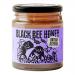 Six glass jars of Black Bee British Autumn Honey sit in a neat row, amber honey catching warm light. Each 227g jar has a black lid and a cream label featuring a stylized black bee and autumnal accents. The jars rest on a wooden surface against a soft neutral background. Soft daylight adds depth too.