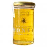 Six clear glass jars contain amber Cambridgeshire summer honey, each topped by a golden lid and labeled Maters & Co Raw Cambridgeshire Summer Honey. The jars stand in two staggered rows on a light surface; warm light glints on the viscous honey and casts soft shadows. Crisp studio styling frames sets.
