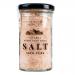 A clear glass jar holds pale pink fine-grain Himalayan salt, packed near the lid. A simple white label wraps the jar with black logo text and minimal graphics. A metal screw cap seals the jar; six identical jars stand grouped on a neutral background. Soft light creates subtle reflection on the glass