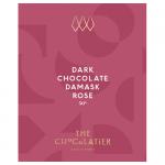 A rectangular dark chocolate bar rests beside a blush-rose wrapper embossed with damask motifs and gold lettering. The chocolate shows glossy squares sprinkled with tiny dried rose petals and a subtle floral sheen beneath soft studio lighting. Subtle shadows and a neutral backdrop emphasize texture.