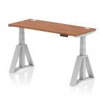 This picture shows a sleek, modern, and versatile dynamic sit-stand height adjustable desk. The desk has a spacious 1400 x 600mm walnut top with cable ports for convenient organization. The silver legs add a touch of elegance, and the piste foot adds stability and support.