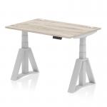 This photograph features a sleek and modern dynamic sit stand and height adjustable desk. The desk measures 1200 x 800mm and has a grey oak top with cable ports. The legs are silver and feature a piste foot to keep the desk stable.