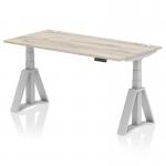 The photograph captures a sleek and modern office desk measuring 1600 x 800mm. Its grey oak top contrasts beautifully with the silver legs and features convenient cable ports for a clutter-free workspace. The desk also boasts a dynamic sit-stand function and a piste foot, perfect for those who value ergonomics and productivity in a stylish package.