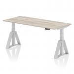 The image shows a modern office desk with a grey oak top and silver legs. It is 1800 x 800mm in size and features cable ports for organization. The desk can be adjusted in height and has a dynamic sit-stand option for improved ergonomics. Its Piste Foot enhances stability and support.